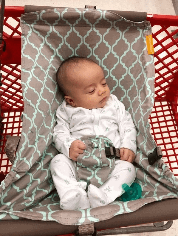 A baby sleeping in a patterned gray and teal hammock-style seat inside a red shopping cart.