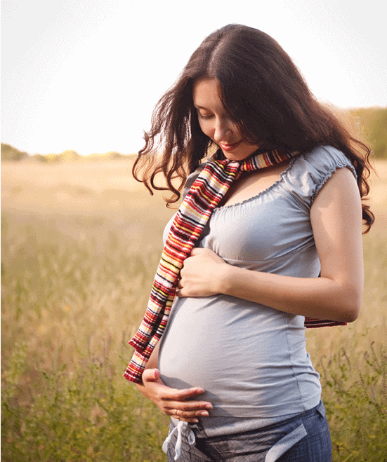 A pregnant woman in a striped scarf cradles her baby bump in a sunlit field.