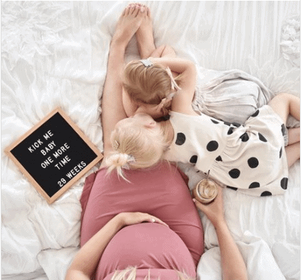 Pregnant woman laying in bed with two young children and a letter board reading KICK ME BABY ONE MORE TIME 29 WEEKS.