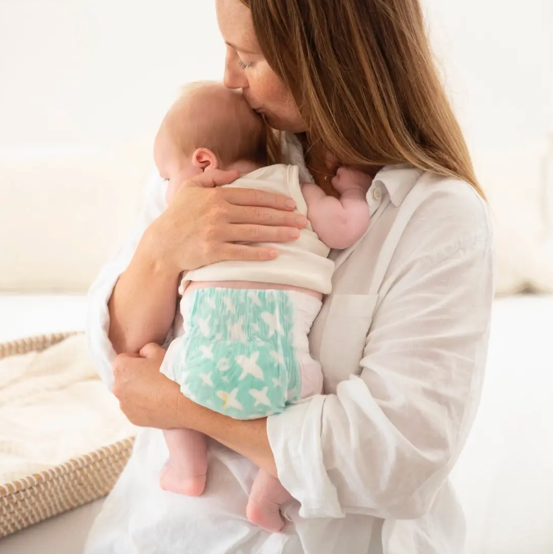 A mother in a white shirt kisses the head of a baby wearing a patterned diaper while holding them against her shoulder.