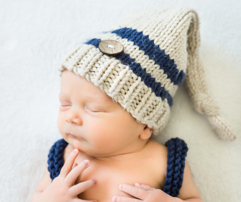 Sleeping newborn wearing a beige and blue striped knit hat with a button and matching knit suspenders.