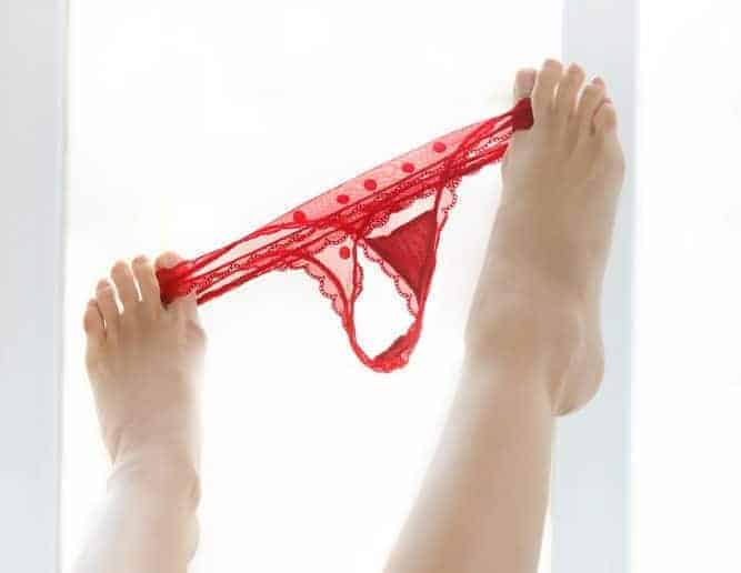 Bare feet hold up a pair of red lace panties against a bright white background, echoing the uncomfortable tone of sex after childbirth and recovery.