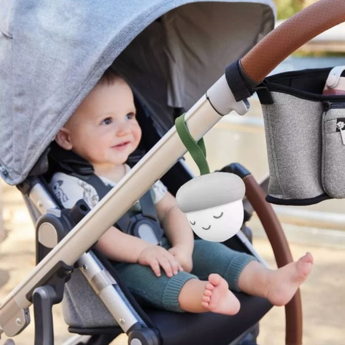 Acorn-shaped portable sound machine with a smiling face hanging from the side of a stroller with a baby inside.