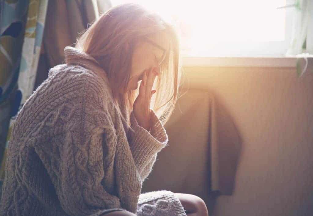 A person wearing a thick knit sweater sits by a bright window and yawns with their hand to their face.
