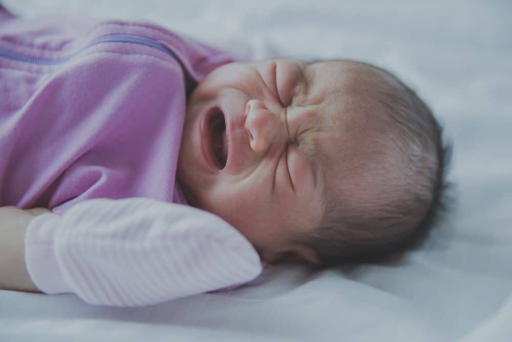 A crying newborn baby wearing a purple sleep sack lies on a white surface with eyes tightly shut and mouth open.