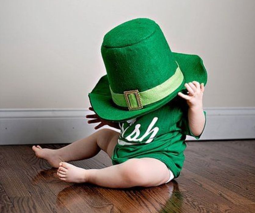 A baby wearing an oversized green leprechaun hat and green t-shirt sitting on a wooden floor.