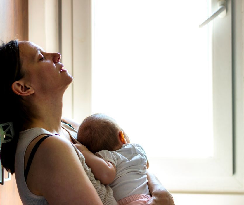 Exhausted mother leaning against a wall with eyes closed, holding a sleeping newborn baby by a bright window.
