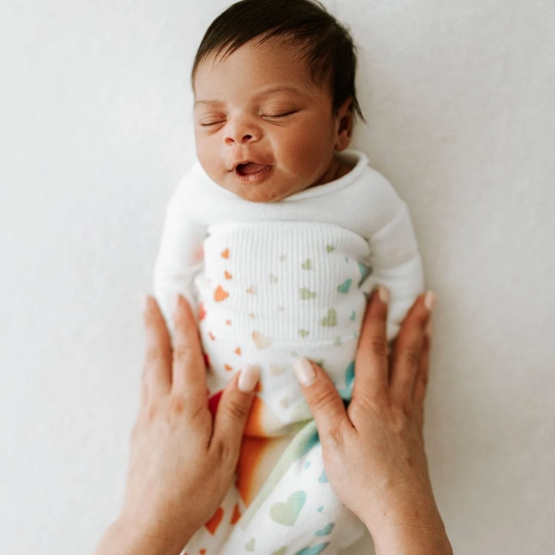 Sleeping infant in a white Swaddelini sleep sack with a rainbow and heart pattern, held by adult hands.