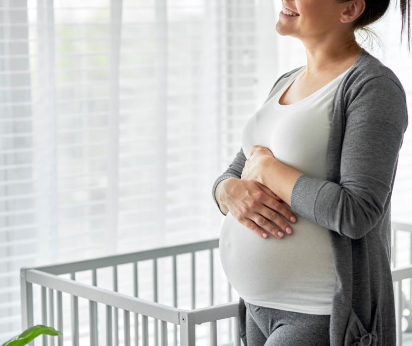 Smiling pregnant woman in a gray cardigan holding her belly while standing next to a white crib.