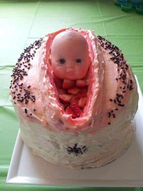 A pink frosted cake with a plastic baby doll head emerging from a vertical opening filled with sliced strawberries.