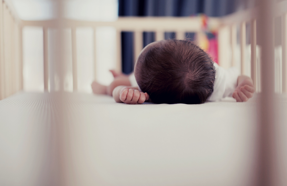 A baby with dark hair sleeping on their stomach in a wooden crib, seen from behind.