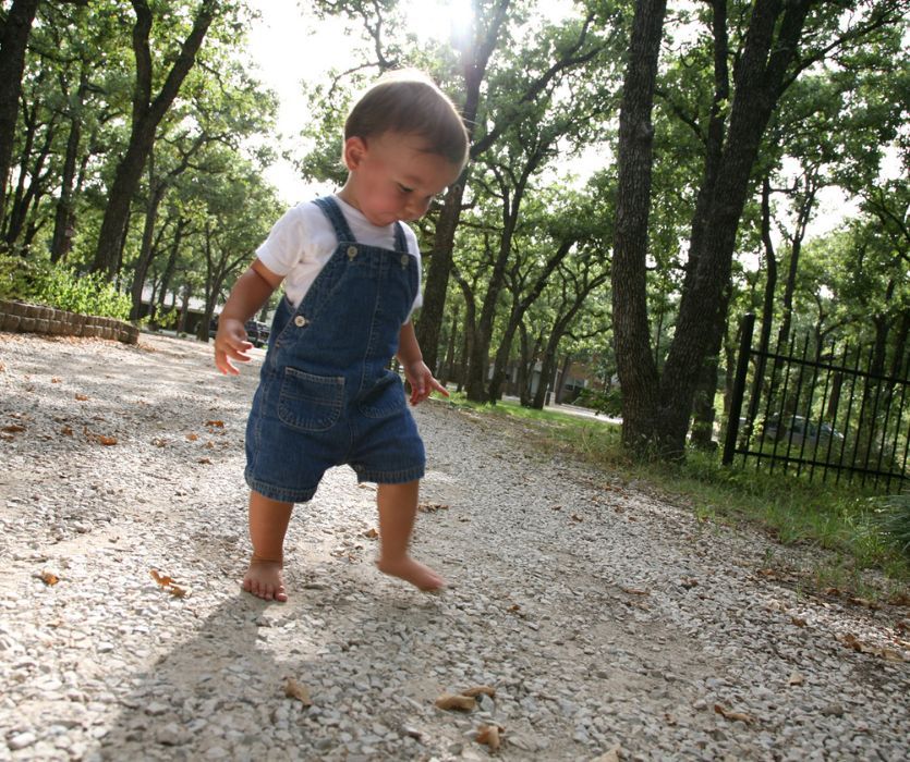 A barefoot toddler in denim overalls walks on a gravel path through a wooded area.