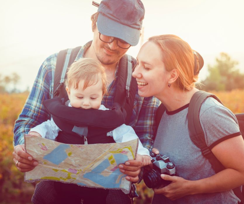 A father carrying a baby in a front carrier and a mother holding a camera look at a paper map while outdoors.