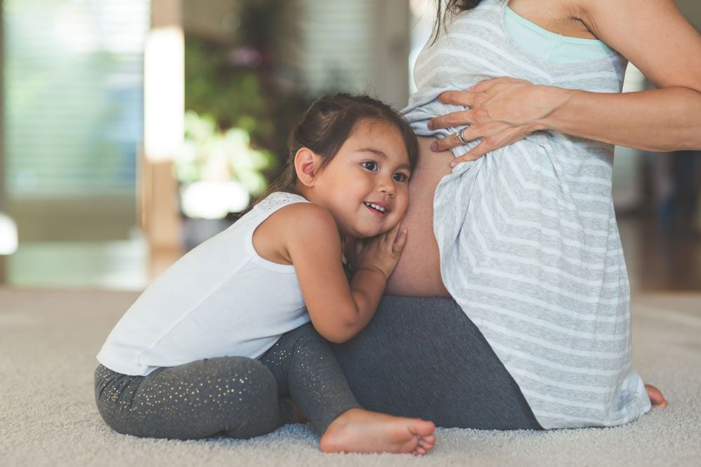 A young girl smiles while leaning her head against a woman's pregnant belly.