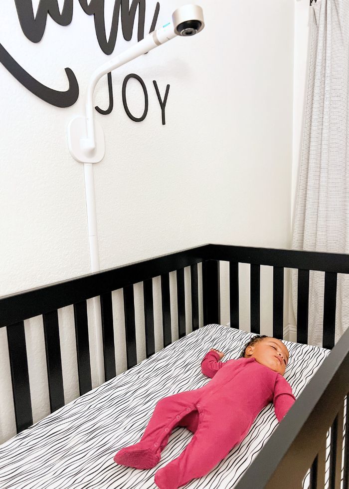 Baby sleeping in a black crib with a VTech V-Care Smart Nursery Camera mounted on the wall above.