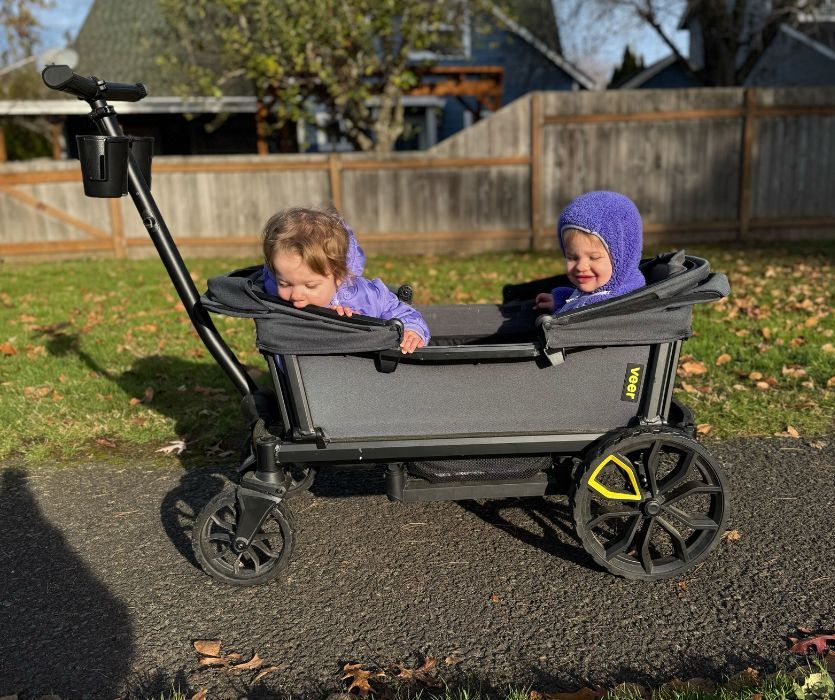 Two young children sitting in a gray Veer Cruiser stroller wagon on a paved path outdoors.