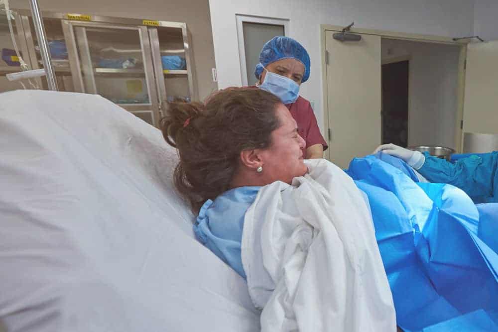A woman in a hospital bed pushes during labor as a healthcare worker in a mask and cap observes.