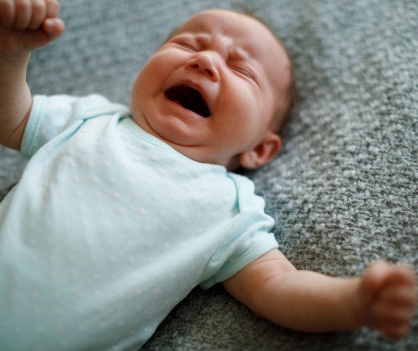An infant in a light blue onesie lying on a gray textured blanket with eyes closed and mouth open, crying.