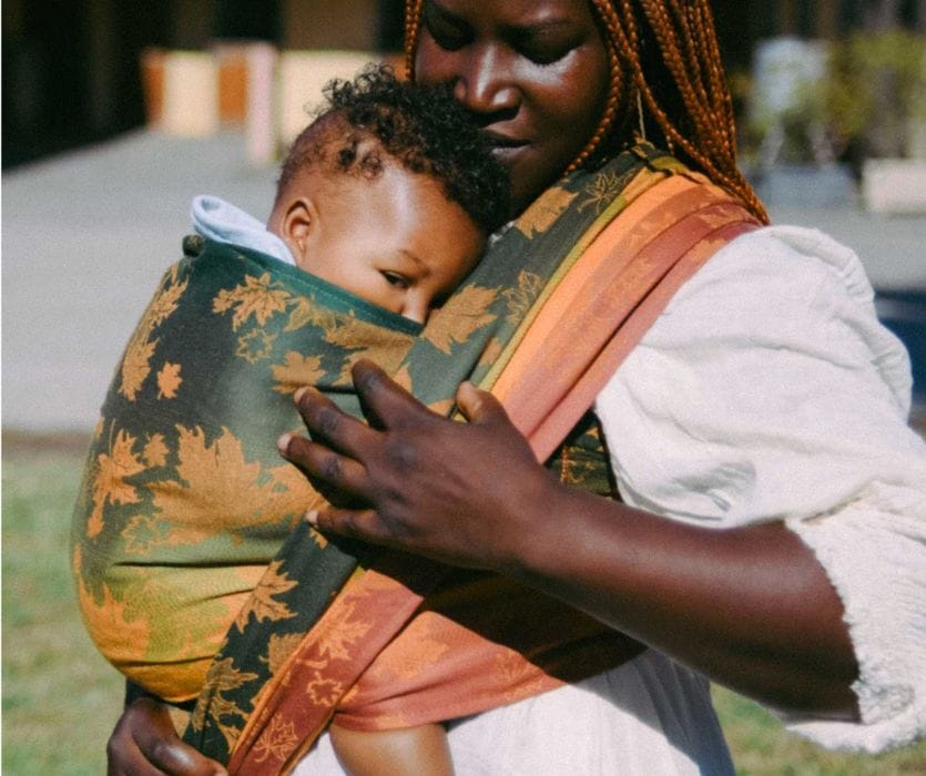 A person carrying a baby in a leaf-patterned woven wrap baby carrier.
