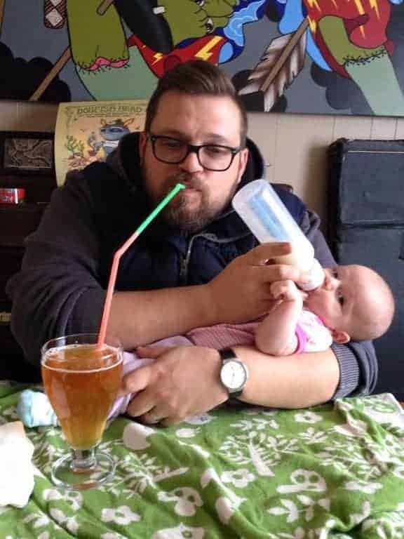 A man drinks a beer through a long straw while bottle-feeding an infant in his arms.