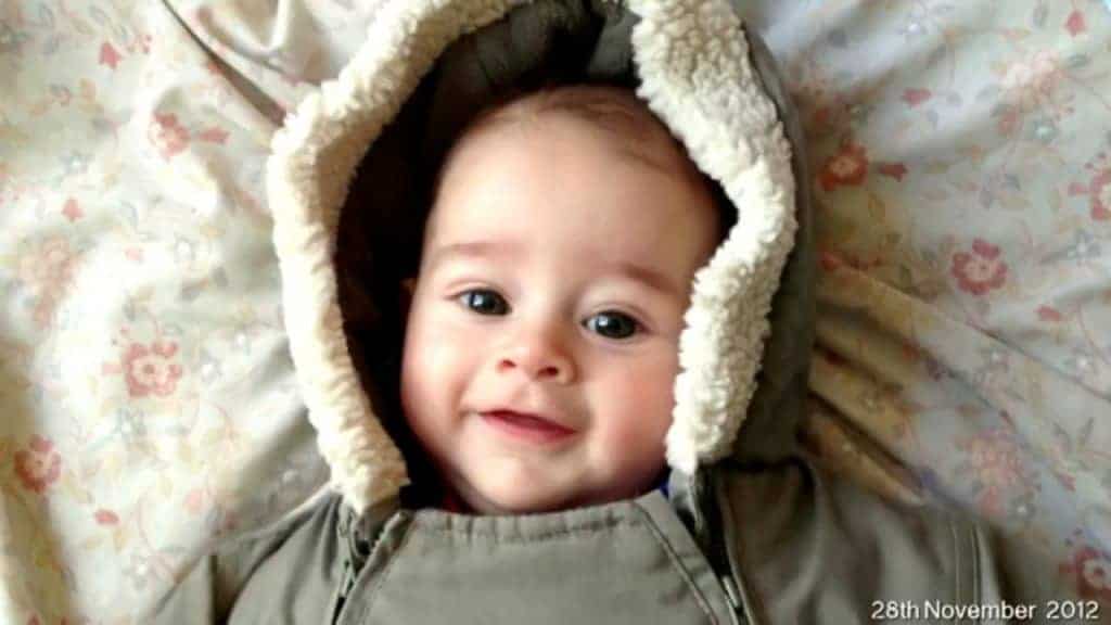 Smiling baby wearing a hooded winter coat lying on a floral blanket. Text at bottom reads 28th November 2012.