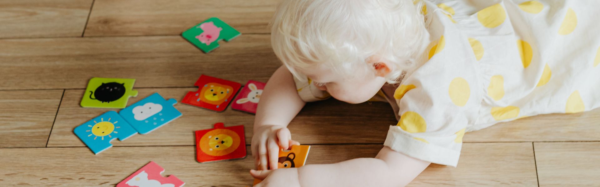 A young child with blonde hair lies on a wooden floor, playing with colorful animal and weather puzzle pieces.