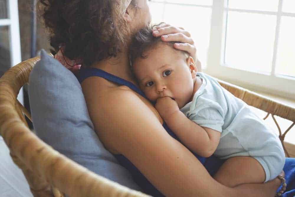A woman sits in a wicker chair holding a toddler who is resting their head on her shoulder while looking at the camera.
