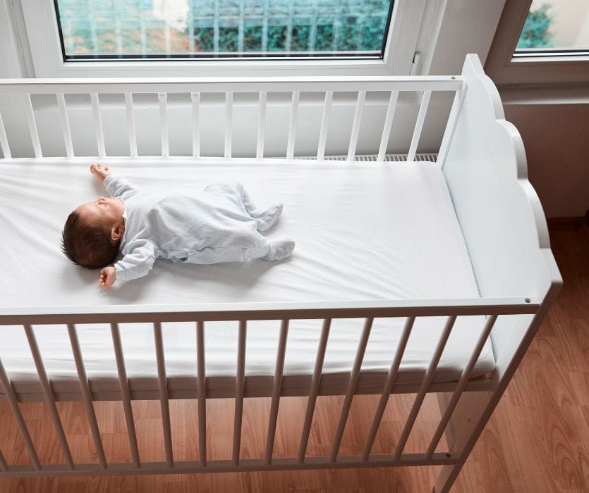 A baby sleeping on their back in a white wooden crib next to a window in a nursery.