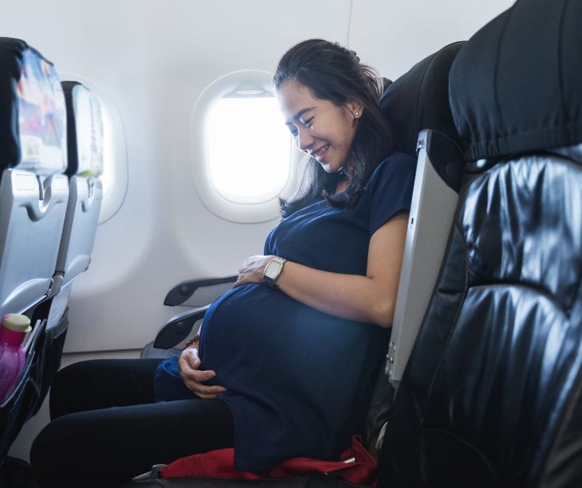 A smiling pregnant woman sits in an airplane seat, cradling her baby bump.