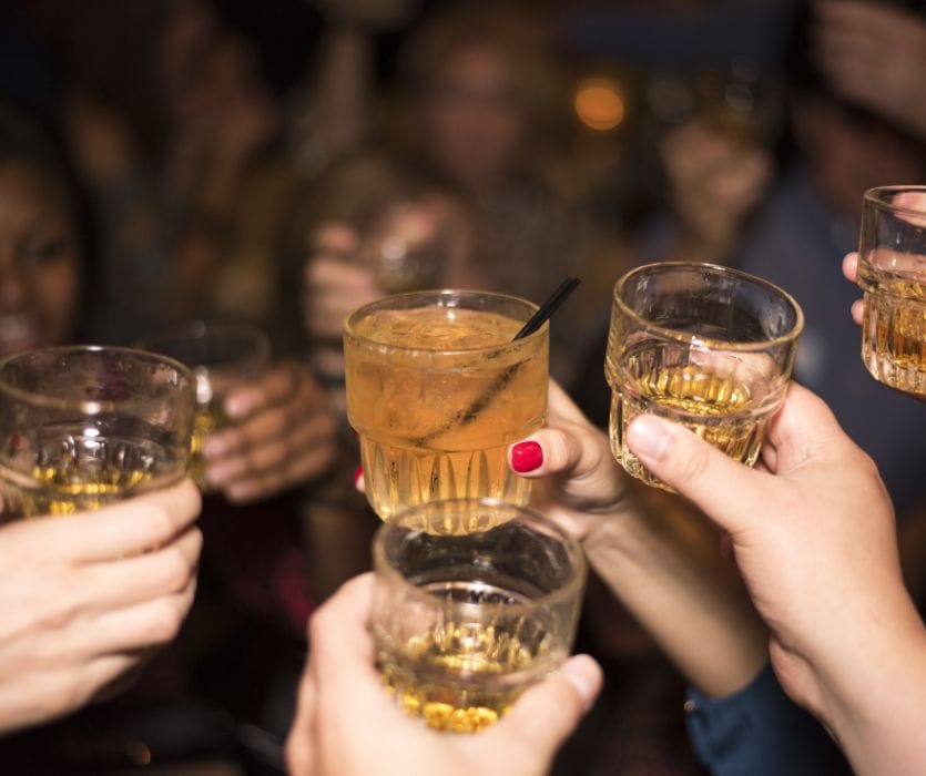 Several hands raise glasses of whiskey and cocktails in a dim party crowd while discussing alcohol and pregnancy safety, with one red-polished hand visible.