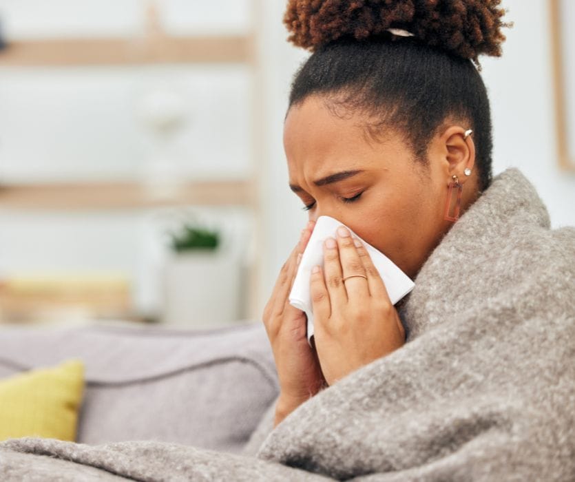 A person with curly hair pulled into a bun blows their nose into a tissue while wrapped in a grey blanket.
