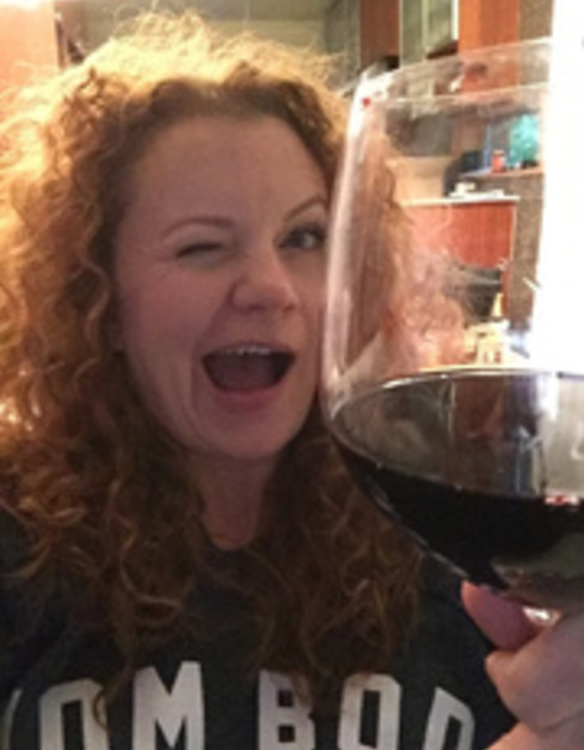Amy winking and smiling behind a large glass of red wine.
