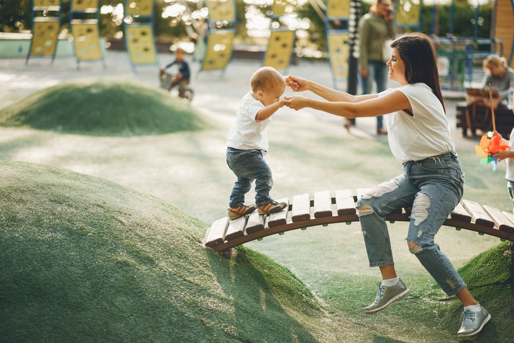 A smiling mother holds hands with her toddler as he walks across a wooden bridge at a sunlit playground.