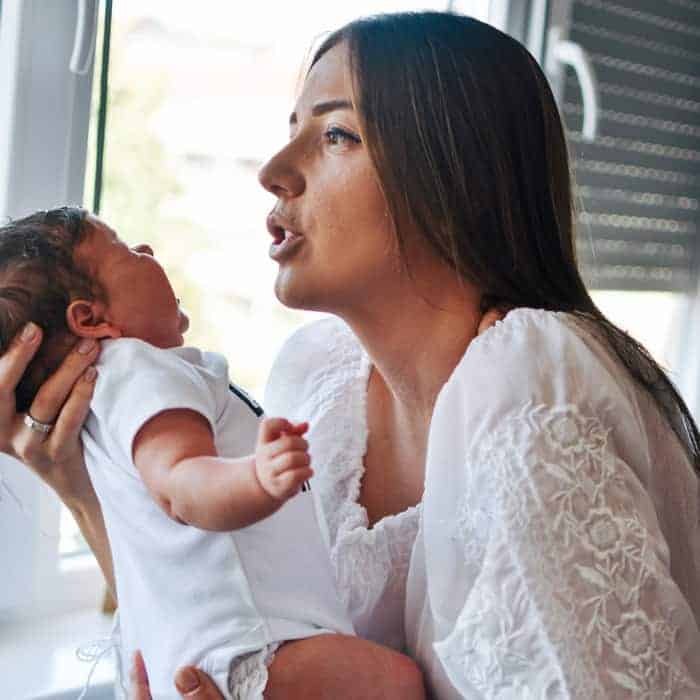 A woman in a white lace shirt holds and looks down at a crying newborn baby in a white onesie.