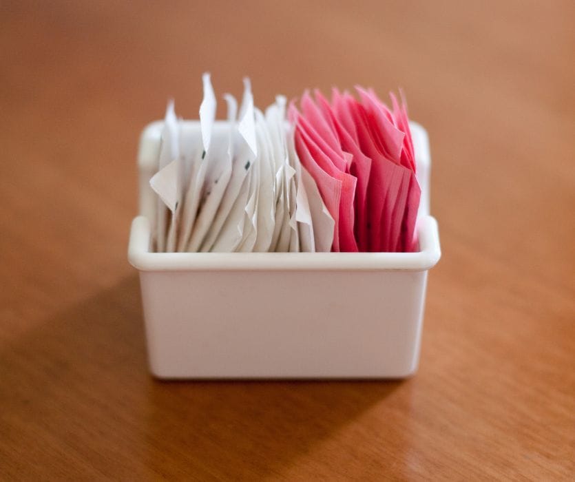 White and pink artificial sweetener packets in a small white container on a wooden table.