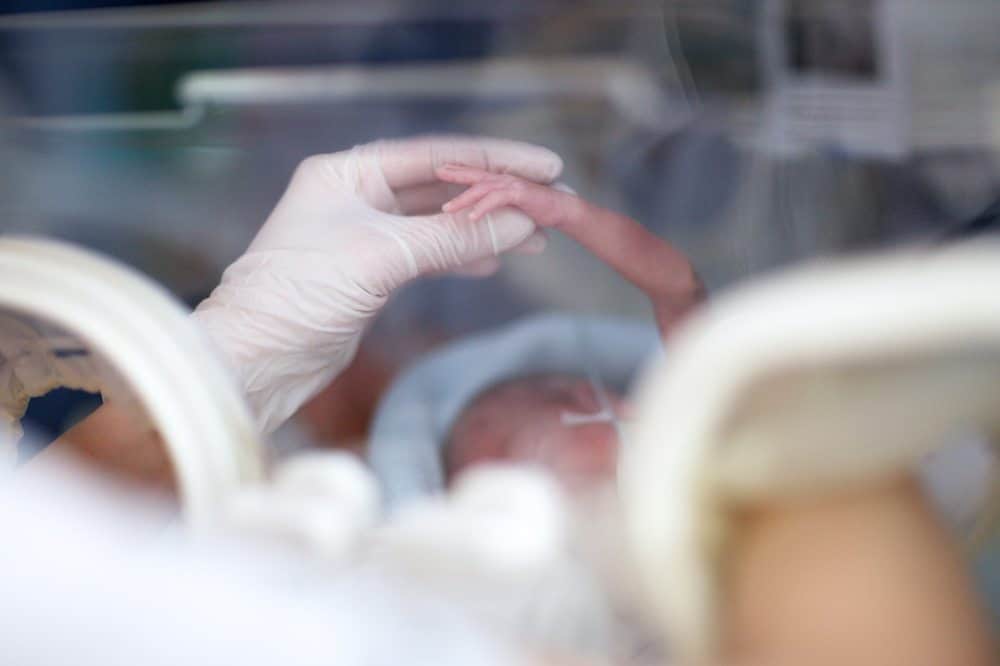A gloved hand gently holding the tiny hand of a premature baby inside a medical incubator.