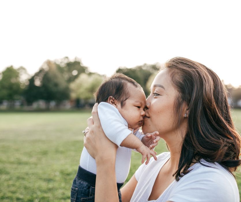 A smiling woman in a park holds and kisses a newborn baby on the nose.