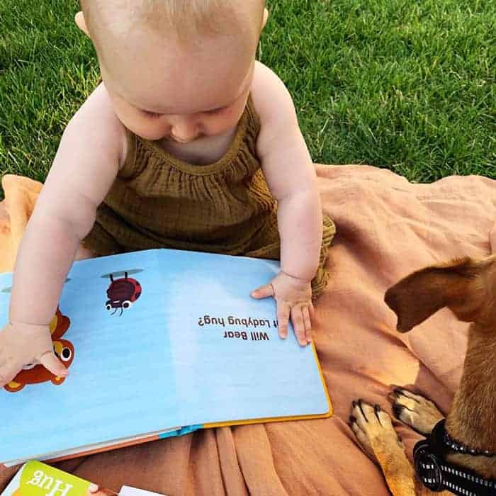 A baby reads a ladybug-themed board book on a blanket outdoors next to a small dog.
