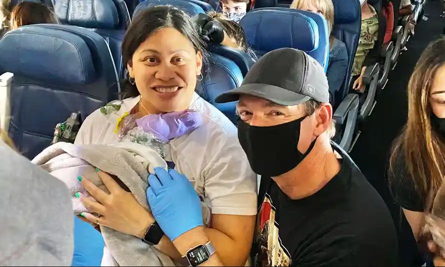 Smiling woman and a man in a face mask holding a newborn baby wrapped in a blanket on an airplane.