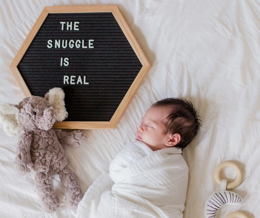 A swaddled newborn sleeping by a plush elephant and a letterboard that reads, The Snuggle Is Real.