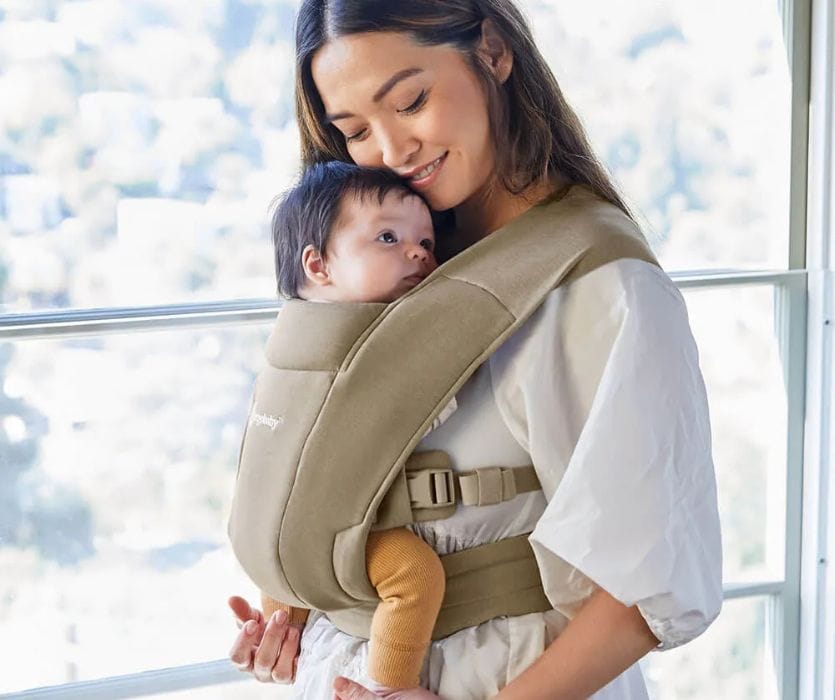 A woman smiles while holding an infant in a tan front-facing baby carrier near a window.