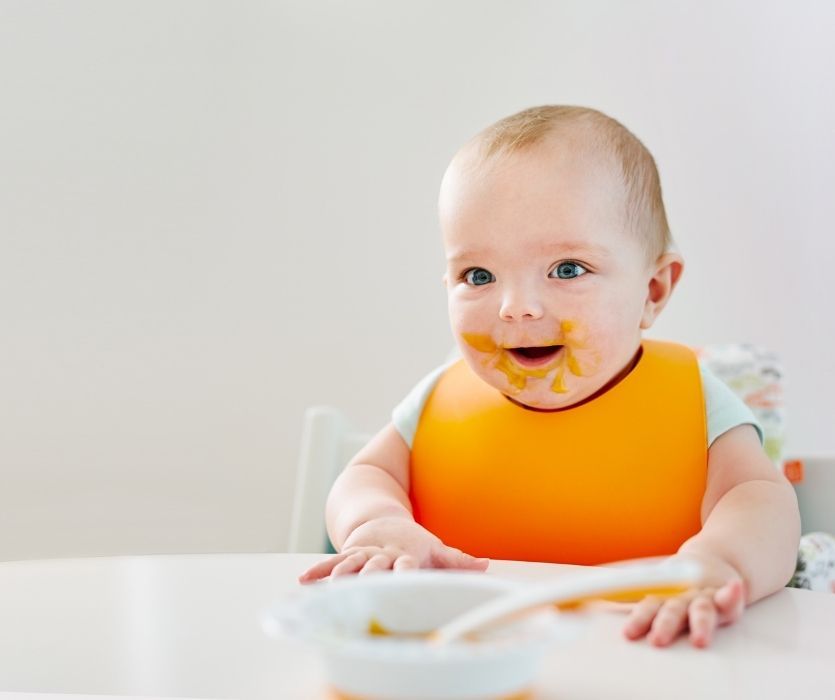 Smiling baby wearing an orange bib with food on their face sits at a white table.