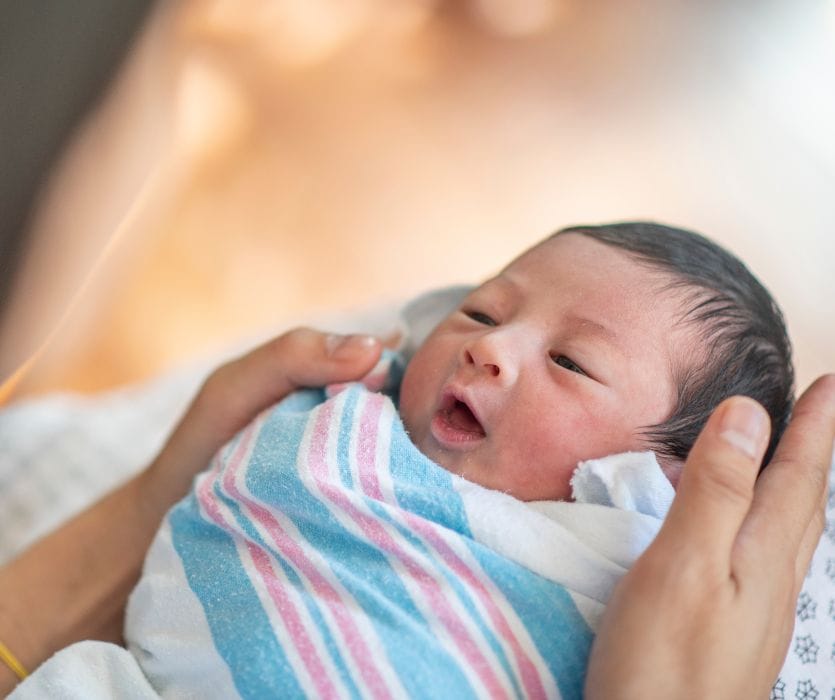 A person cradle-holding a newborn baby swaddled in a blue and pink striped hospital blanket.