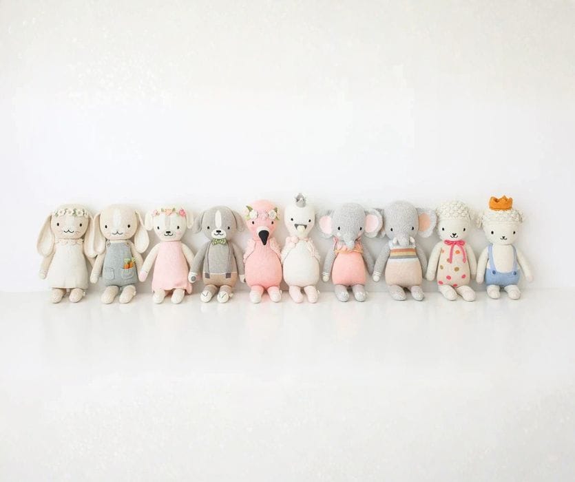 A row of ten different hand-knit animal dolls in pastel clothing against a white background.