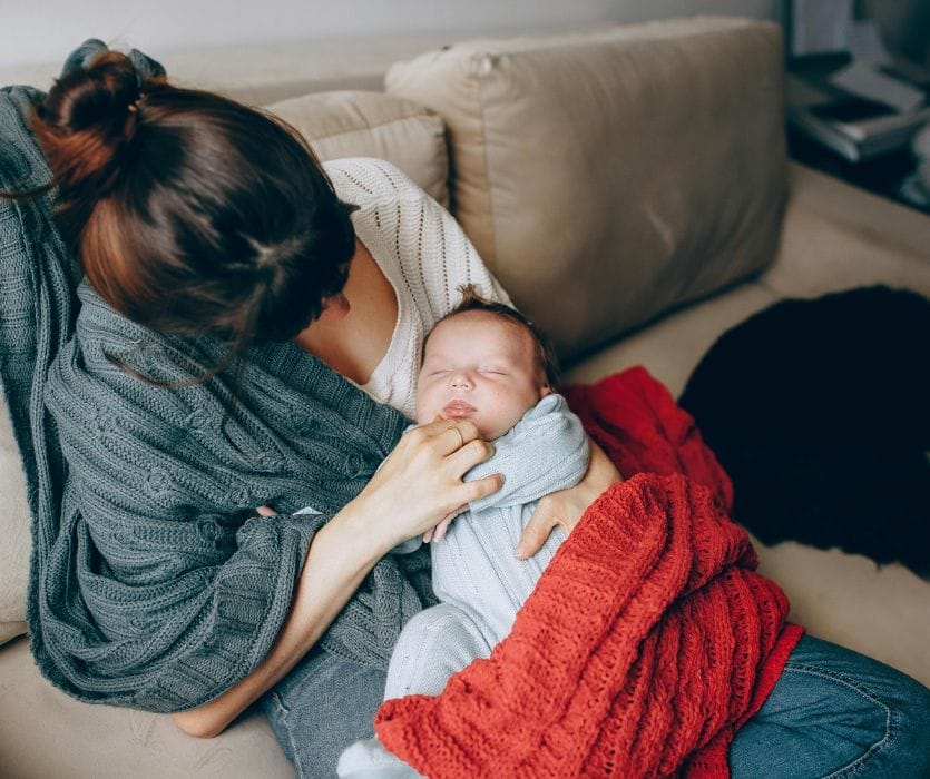 A woman cradles a sleeping newborn baby wrapped in a gray knit sweater and a red textured blanket on a sofa.
