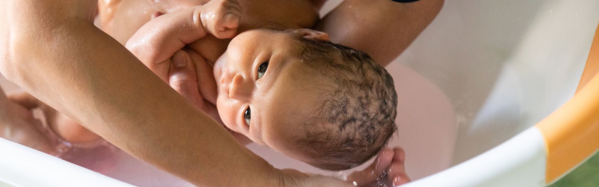A person’s arms supporting and bathing a newborn baby in a small white and orange plastic tub.