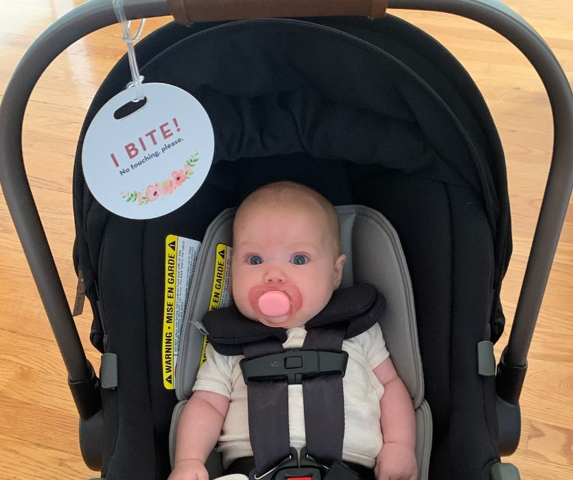 Baby in a car seat with a pacifier and a hanging sign that says, I BITE! No touching, please.