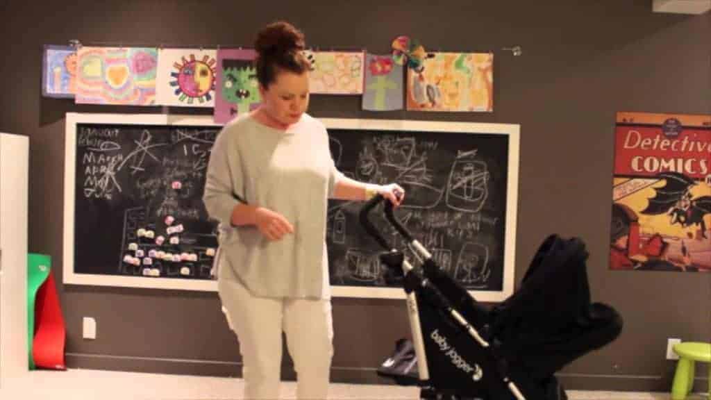 A woman stands next to a black Baby Jogger Vue stroller in a room with a chalkboard and children's artwork.