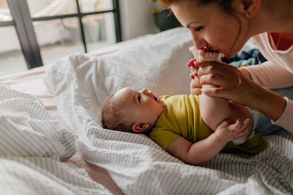 A mother plays with her laughing baby on a bed, gently holding and kissing the infant's feet.