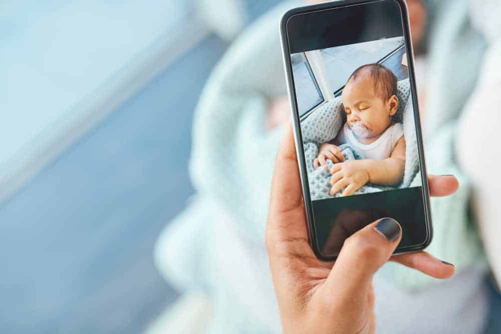 A person holding a smartphone displaying a photo of a sleeping newborn baby wrapped in a blue blanket.